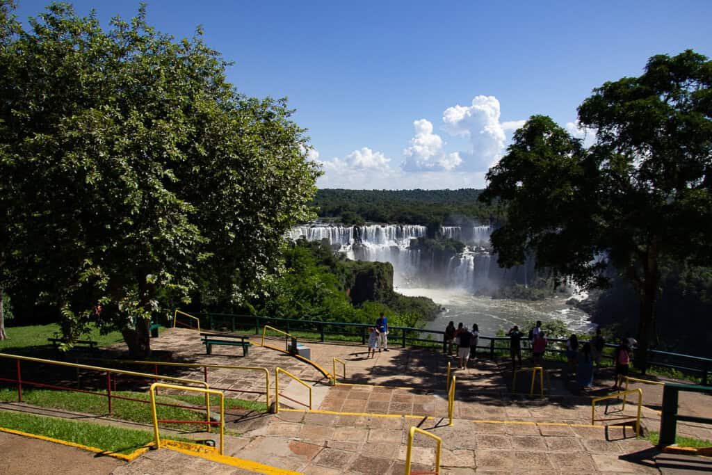 The image captures a vibrant scene at the start of a trail on the Brazilian side of Iguazu Falls. A group of visitors is gathered at a viewpoint, overlooking the spectacular waterfalls in the distance. The falls are a series of vast and powerful cascades, creating a dramatic backdrop against the clear sky, with a few puffy clouds floating above. Lush greenery frames the scene, with a large tree in the foreground providing shade and a sense of scale. The observation area is marked by yellow railings and paved with stone tiles, suggesting an accessible path for tourists to enjoy the natural beauty of the area. The photo conveys a sense of wonder and the allure of exploring one of the world's most magnificent waterfalls.