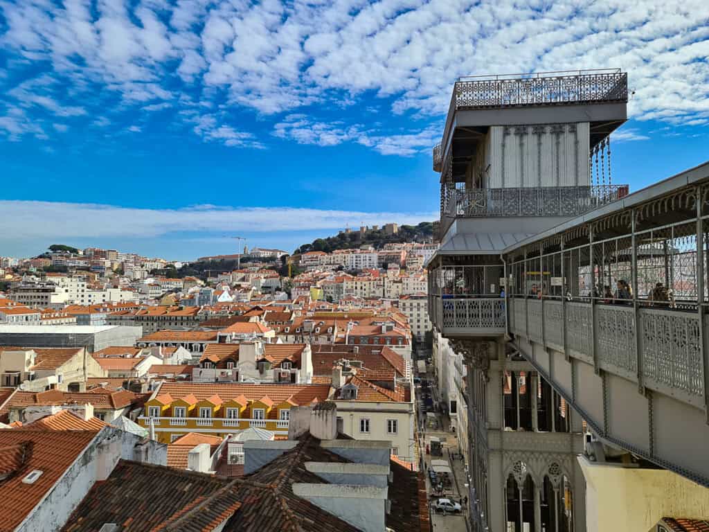 A panoramic view from the Santa Justa Lift overlooking the rooftops of Lisbon. The lift's intricate metalwork is in the foreground, with the city's colorful buildings, hills, and a clear sky in the background.
