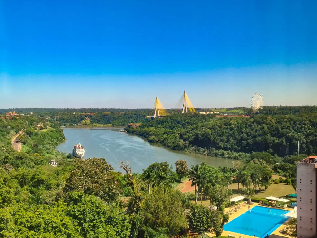 The photograph displays a scenic view from the hotel room in Puerto Iguazu, showcasing a serene river flanked by lush greenery. A prominent bridge with distinctive yellow triangular structures spans the river in the distance. On the right, a large Ferris wheel creates a focal point against the clear sky. In the foreground, a swimming pool with azure water provides a leisurely contrast to the natural scene.