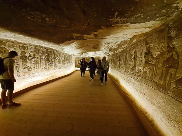 A group of tourists exploring an ancient, dimly-lit Egyptian tomb with intricately carved hieroglyphs on the walls.