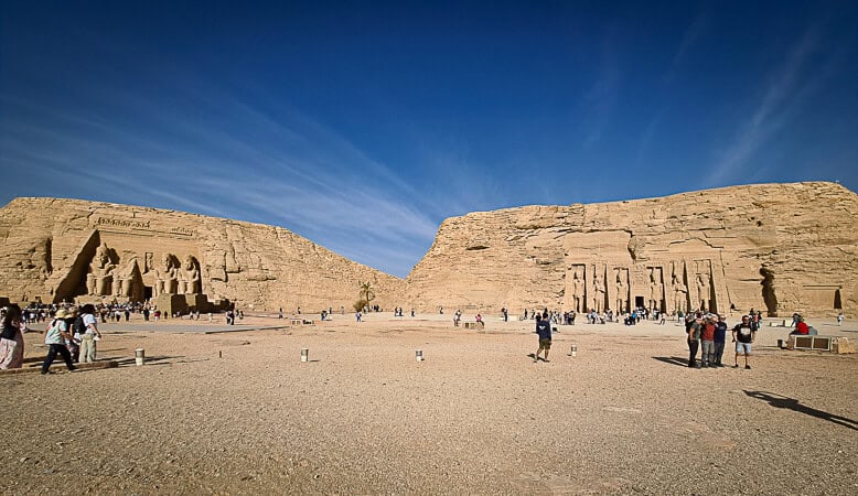 This image shows a wide-angle view of a historic Egyptian monument, the Temple of Ramses II at Abu Simbel. The temple's facade is adorned with four colossal statues of Ramses II seated on thrones, each several meters high, with smaller figures at their feet representing family members. The temple is carved directly into a solid rock cliff and the entrance is between the statues. There's a clear blue sky above and a broad, sandy area in front of the temple where visitors are scattered around, some taking photos, others walking towards the temple. The scale of the human figures compared to the statues and the temple itself emphasizes the monument's grandeur.