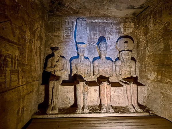 This image depicts an interior view of an ancient Egyptian temple. It shows a dimly lit sanctuary with four large seated statues carved into the rock face. The statues are likely representations of deities or pharaohs, as indicated by their traditional crowns and the uraeus (cobra) on the forehead of two figures. Hieroglyphics and bas-relief carvings adorn the walls surrounding the statues, adding to the historical and cultural significance of the chamber. The lighting casts dramatic shadows, highlighting the depth of the carvings and the texture of the stone.