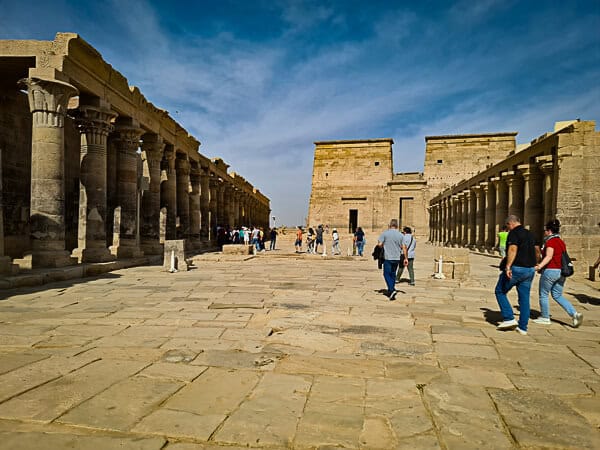 This image shows a bright, sunny day at the Philae Temple complex in Egypt. A paved walkway leads visitors through the site, with rows of pillars on one side and the temple's stone walls on the other, featuring carvings and hieroglyphs. The columns are capped with intricate capitals, some resembling the papyrus plant. Tourists are walking along the path, exploring the ancient site, and the clear blue sky above suggests a warm and clear day. The architecture and the clear weather create a vivid and striking scene that is typical of the historic sites along the Nile River.