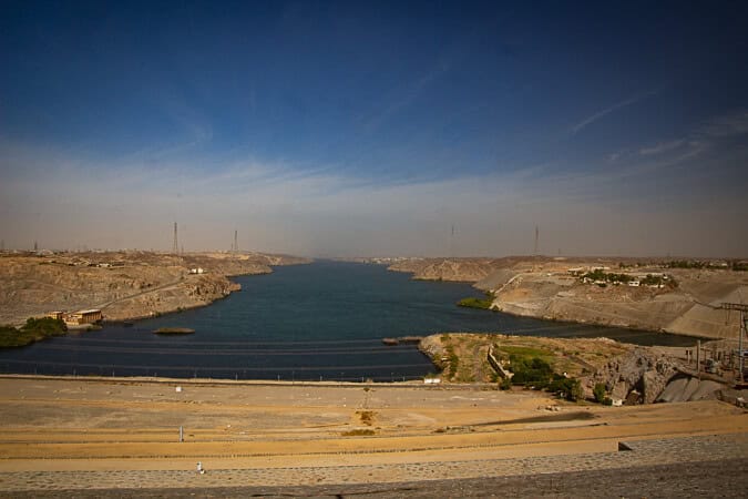 This image provides a broad view of the Aswan High Dam in Egypt, a significant engineering feat on the Nile River. The dam's infrastructure creates a stark contrast against the natural landscape, with the calm blue waters of Lake Nasser in the background. The sky is vast and clear, highlighting the contrast between the arid land and the life-giving river. Electrical transmission lines parallel to the riverbank stretch into the distance, signifying the dam's role in electricity production. The photograph appears to be taken from a high vantage point, offering a sweeping perspective of this monumental structure and its surroundings.