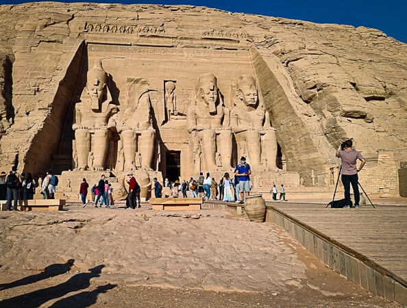 The image shows the Great Temple of Ramses II at Abu Simbel, Egypt, under the clear blue sky. The temple's facade is dominated by four colossal seated statues of the pharaoh, with smaller figures at their feet. Visitors cluster at the entrance, providing a sense of scale to the massive size of the monument. On the right, an individual with a tripod and camera is set up, capturing the scene. The sandstone cliffs that house the temple catch the bright sunlight, emphasizing the impressive artistry and grandeur of this ancient structure.