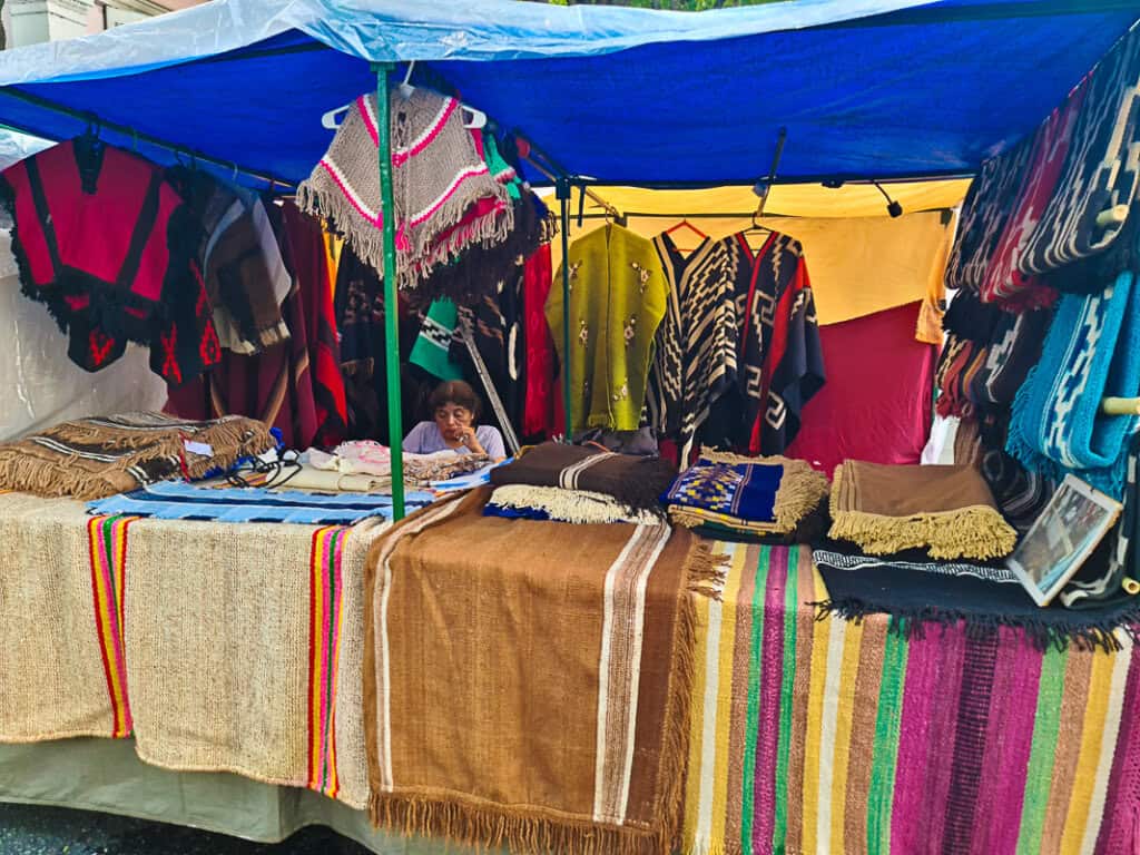 A market stall draped with handwoven textiles exhibits a colorful selection of traditional ponchos and shawls. The various patterns and colors on display reflect the artisan's skill and the cultural heritage of the region. On the table in the foreground, smaller items such as woven bags and handicrafts are presented, while the vendor sits behind, contributing to the authentic atmosphere of the market. The textiles, with their detailed fringes and vibrant stripes, are a testament to the craftsmanship that the Feria de Mataderos is known for.