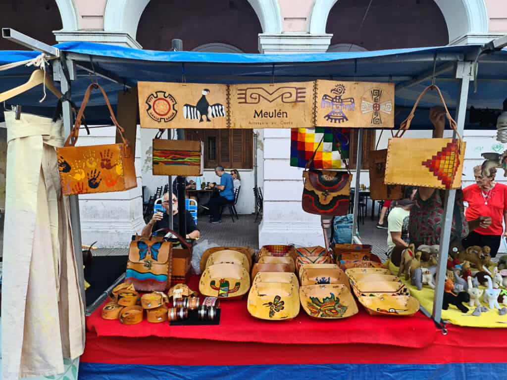 A market stall showcasing traditional Argentine leather crafts, including hand-painted bags, trays, and belts with colorful designs inspired by local culture. Perfect souvenirs from Argentina for lovers of artisan craftsmanship.