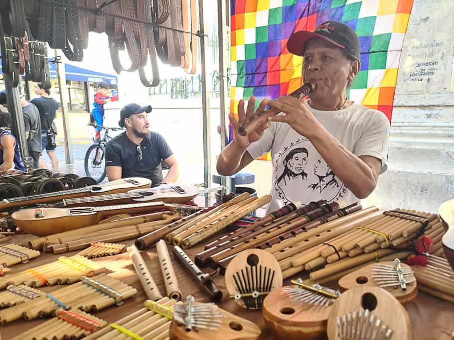 A vendor at a street market in Buenos Aires playing a traditional flute, surrounded by handcrafted musical instruments like pan flutes and charangos. A perfect spot to find authentic Argentinean souvenirs and Argentina gifts reflecting the country's cultural heritage.
