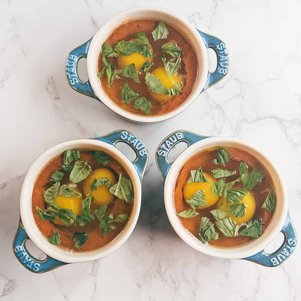 3 mini cocotte dishes with eggs, tomato sauce, and fresh basil.