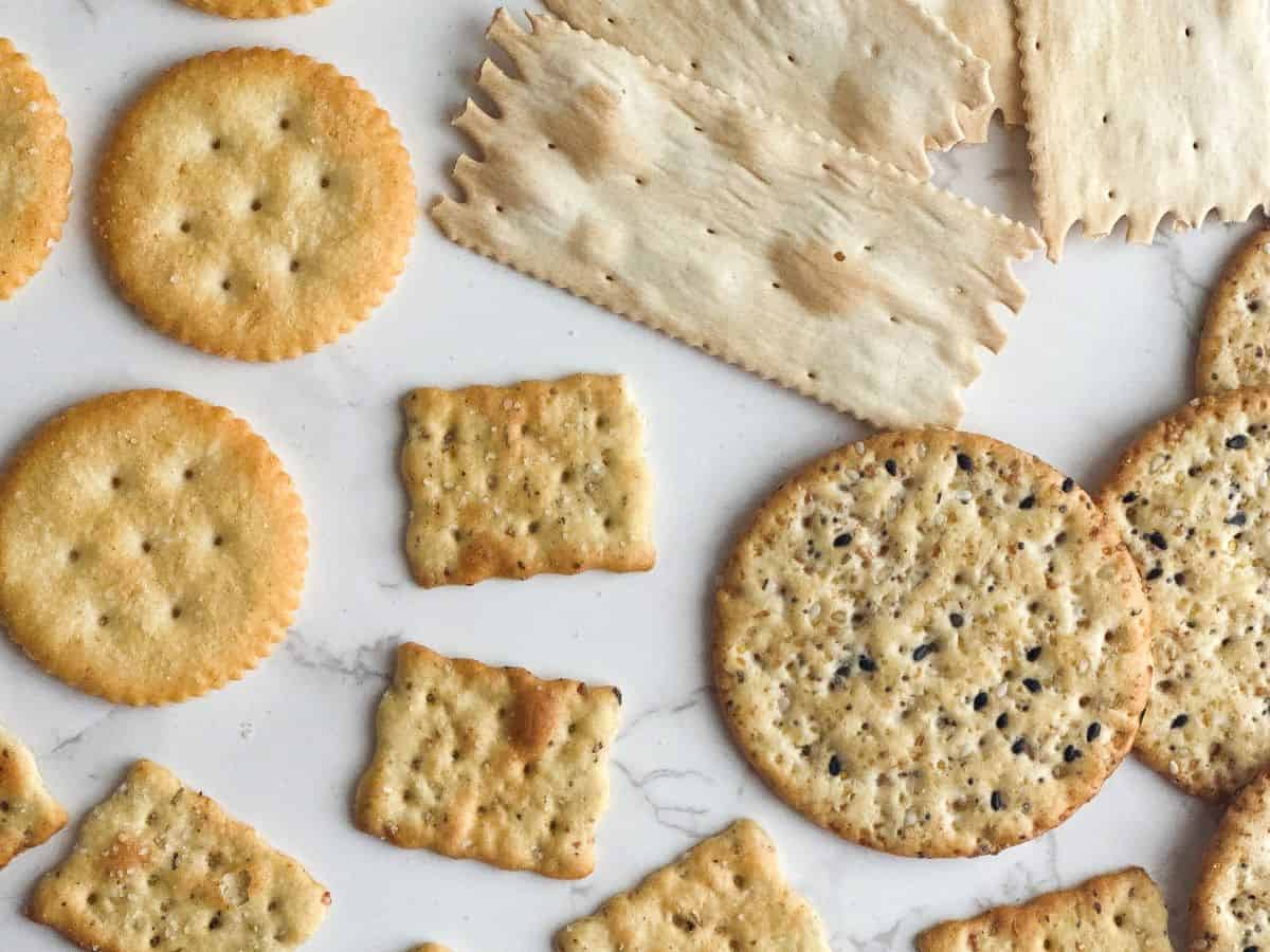 Buttery crackers, multigrain crackers, herb crackers, and flat bread crackers on white marble table top.