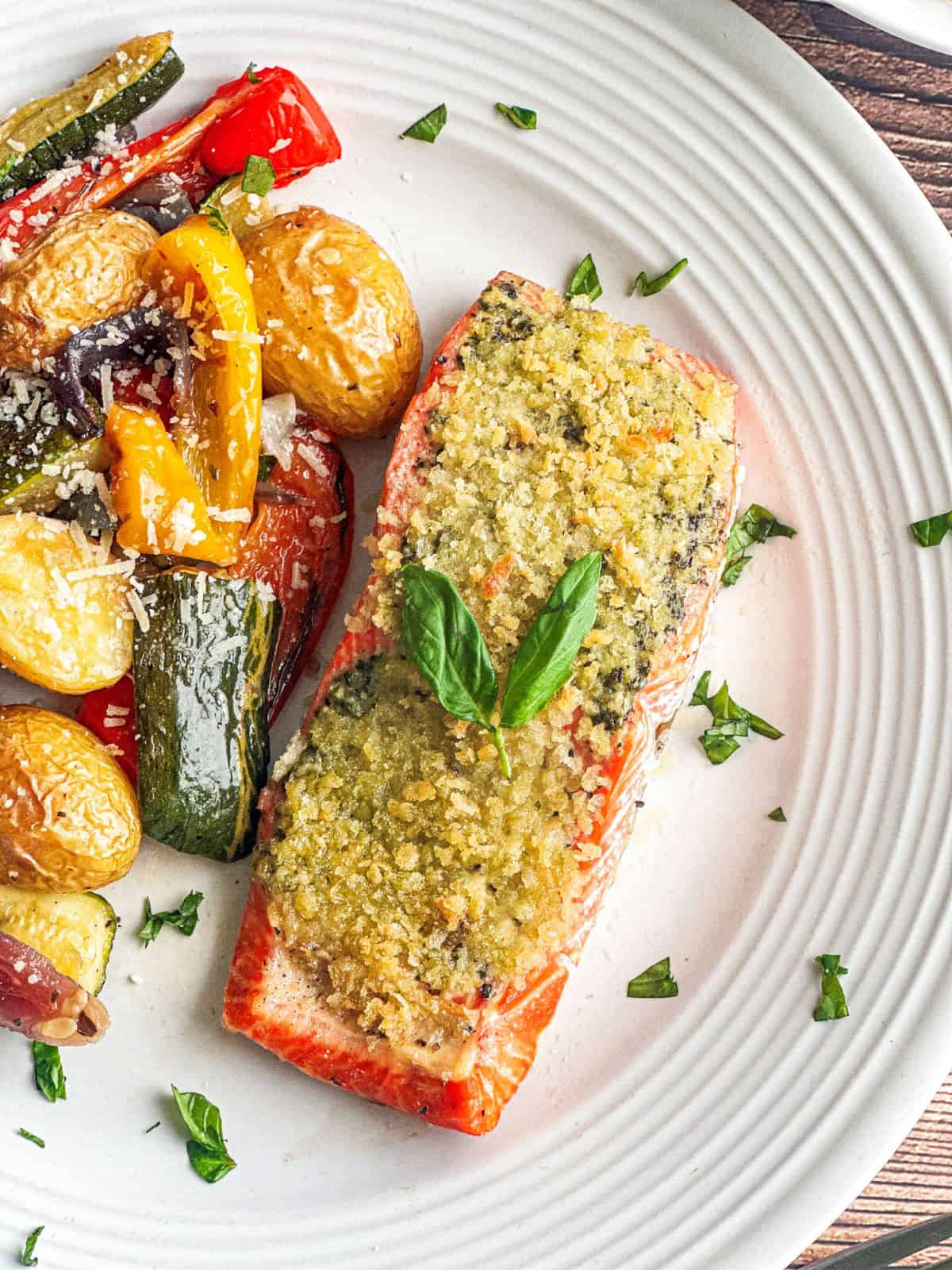 Filet of salmon with pesto crust on a white plate with Italian roasted vegetables.