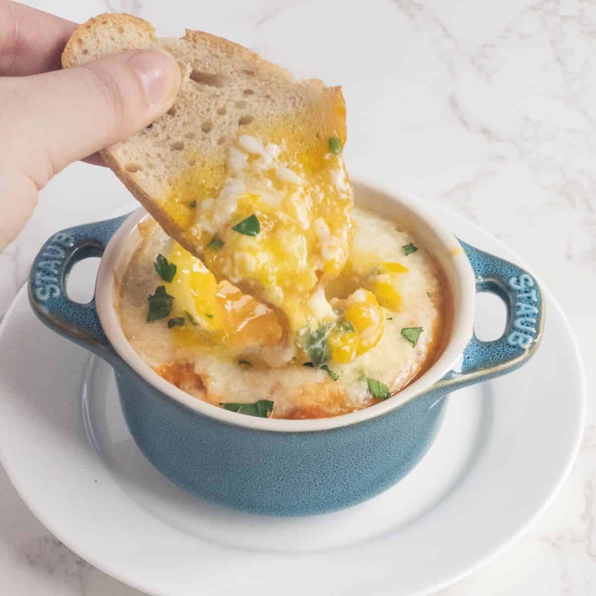 A hand dipping bread into baked eggs in a small cocotte dish.