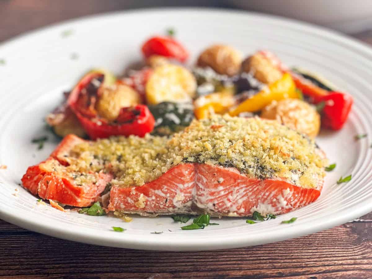 Filet of pesto crusted salmon in a side view on a white plate with vegetables blurred in the background.