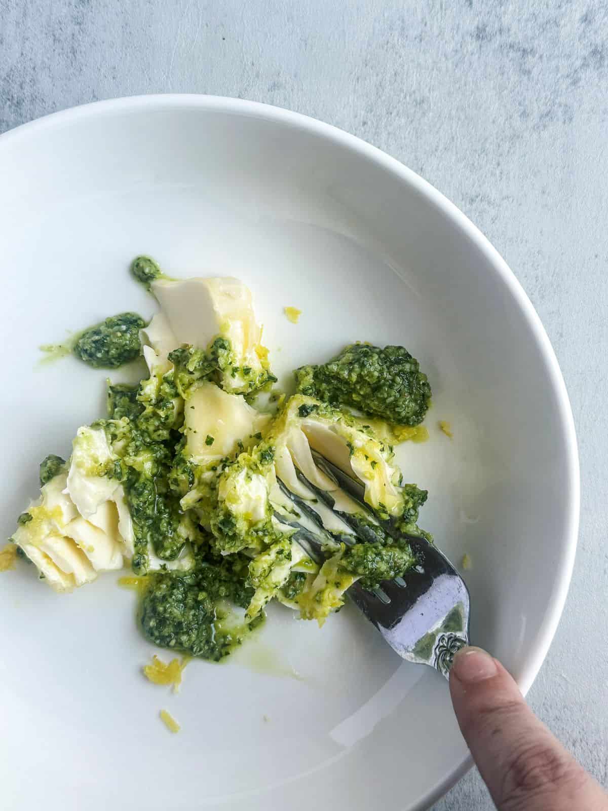 A fork mashing butter and pesto in a white bowl.