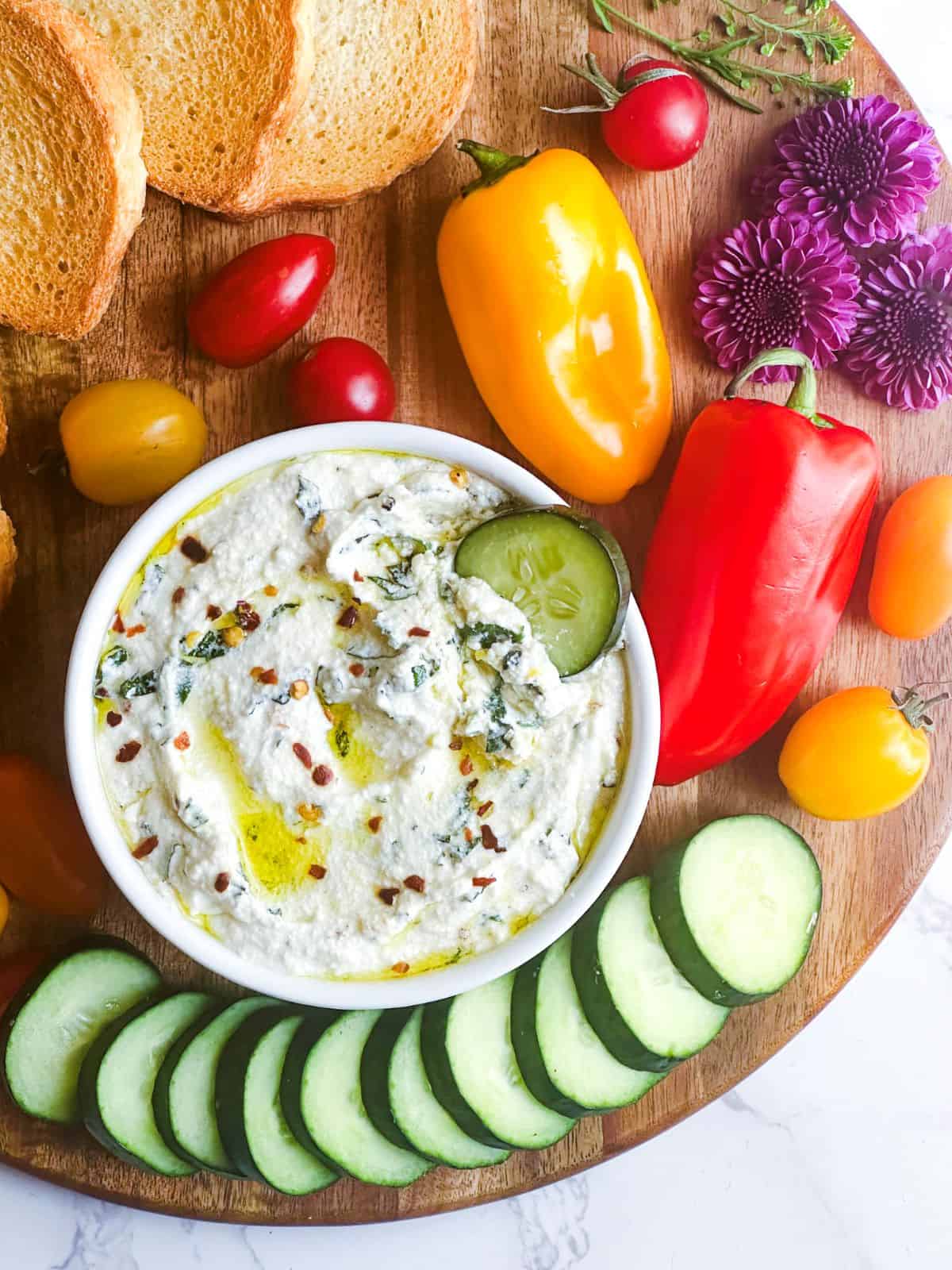 Whipped ricotta dip in a ramekin on a wooden charcuterie board with tomatoes peppers and crostini.