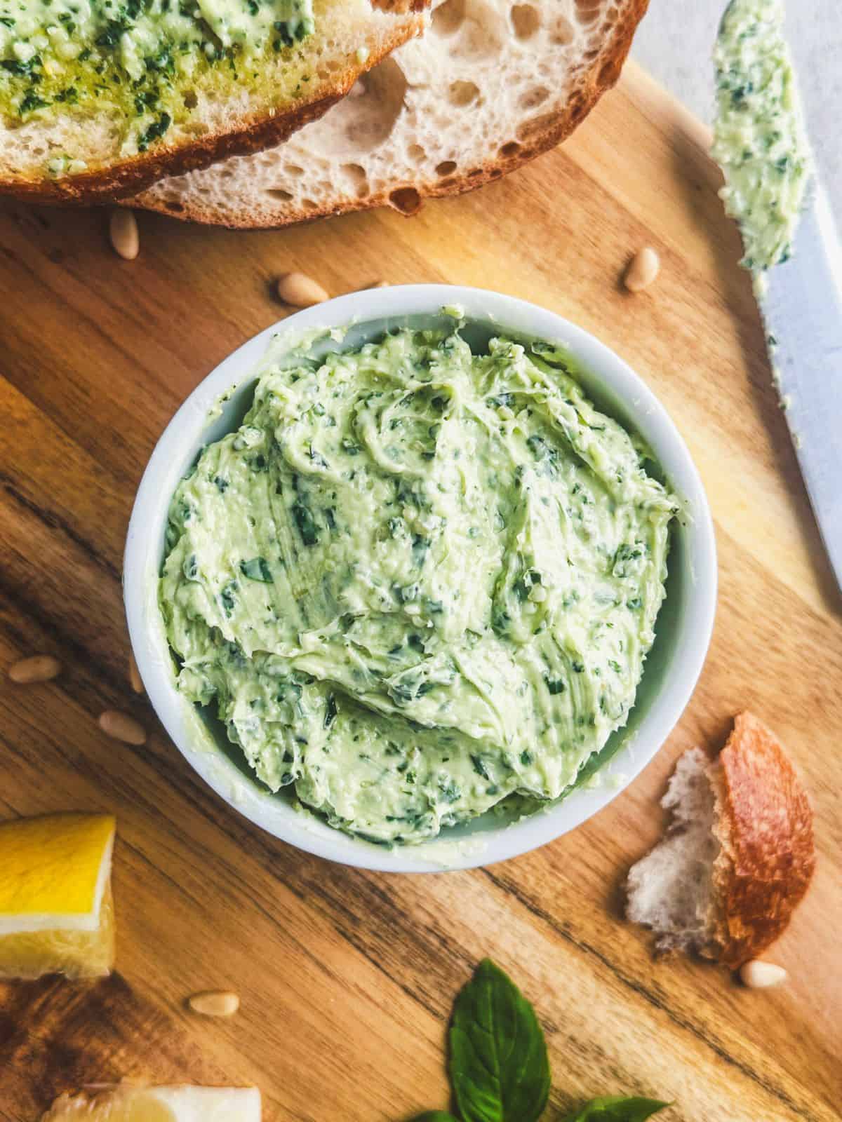 Basil pesto butter in a white ramekin on a cutting board with lemon, fresh basil leaves, pine nuts, and sliced bread.