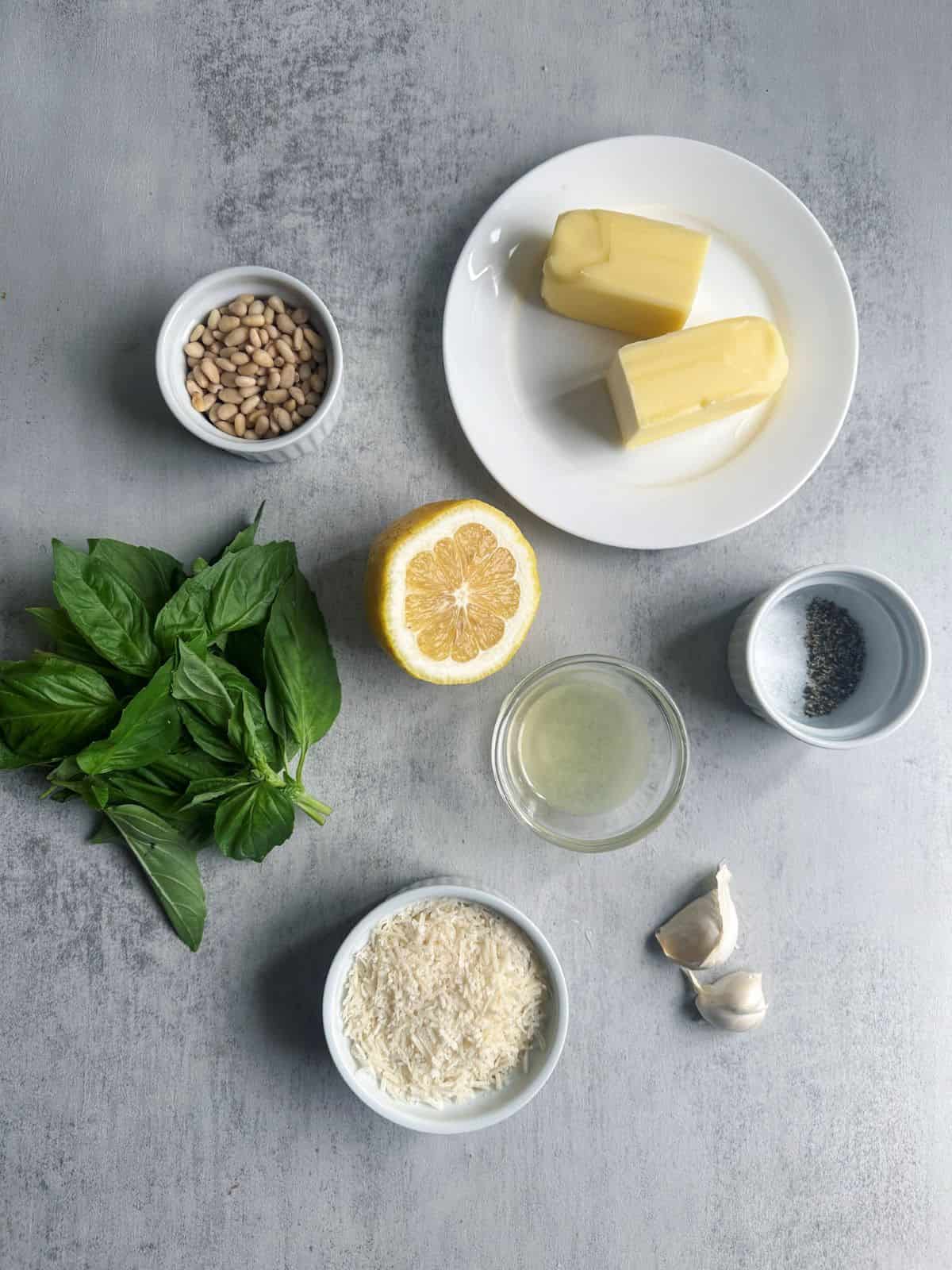 Fresh basil, butter, garlic, parmesan cheese, lemon juice, pine nuts, salt and pepper on a gray background.