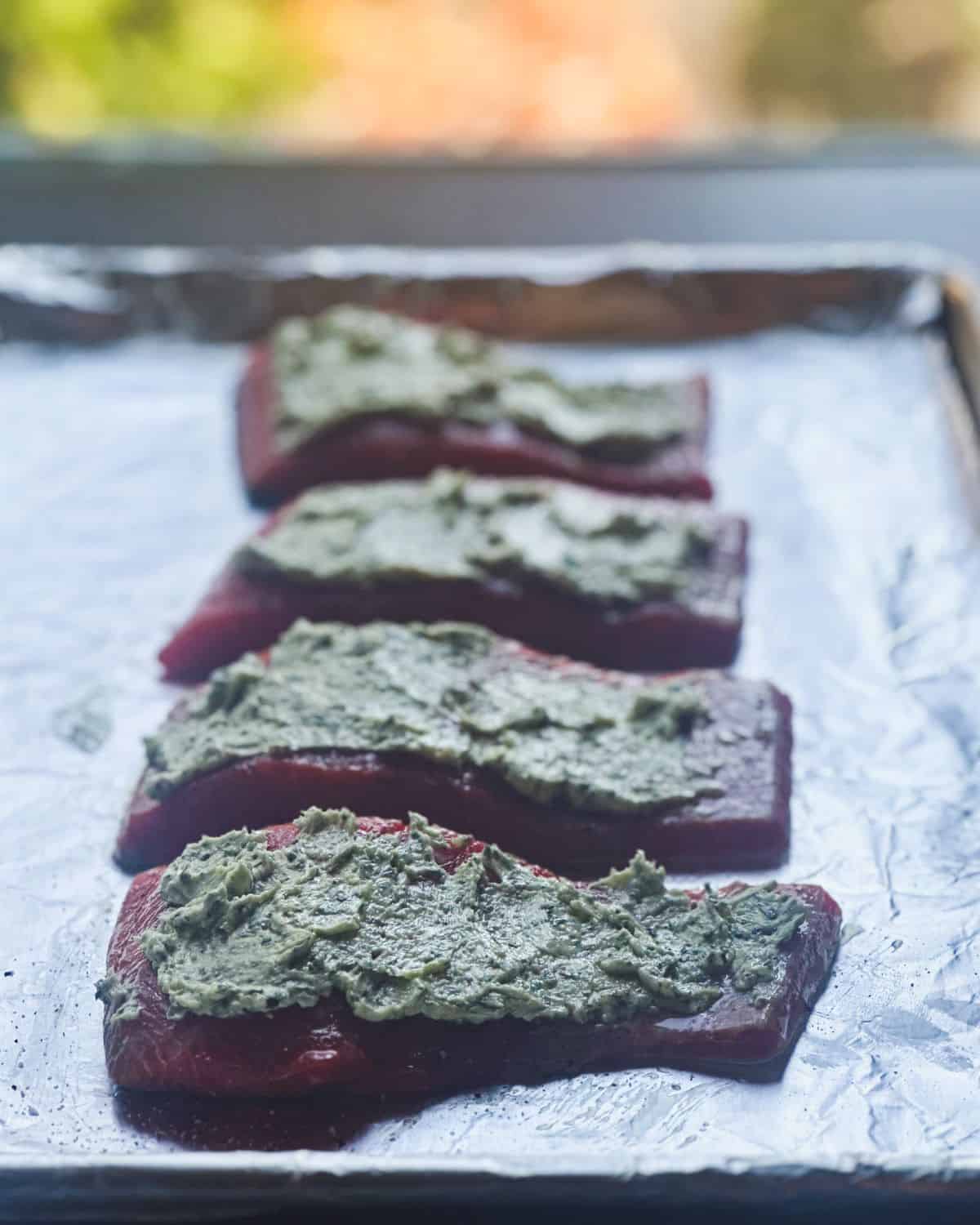 Four uncooked salmon fillets topped with pesto butter on a baking sheet.