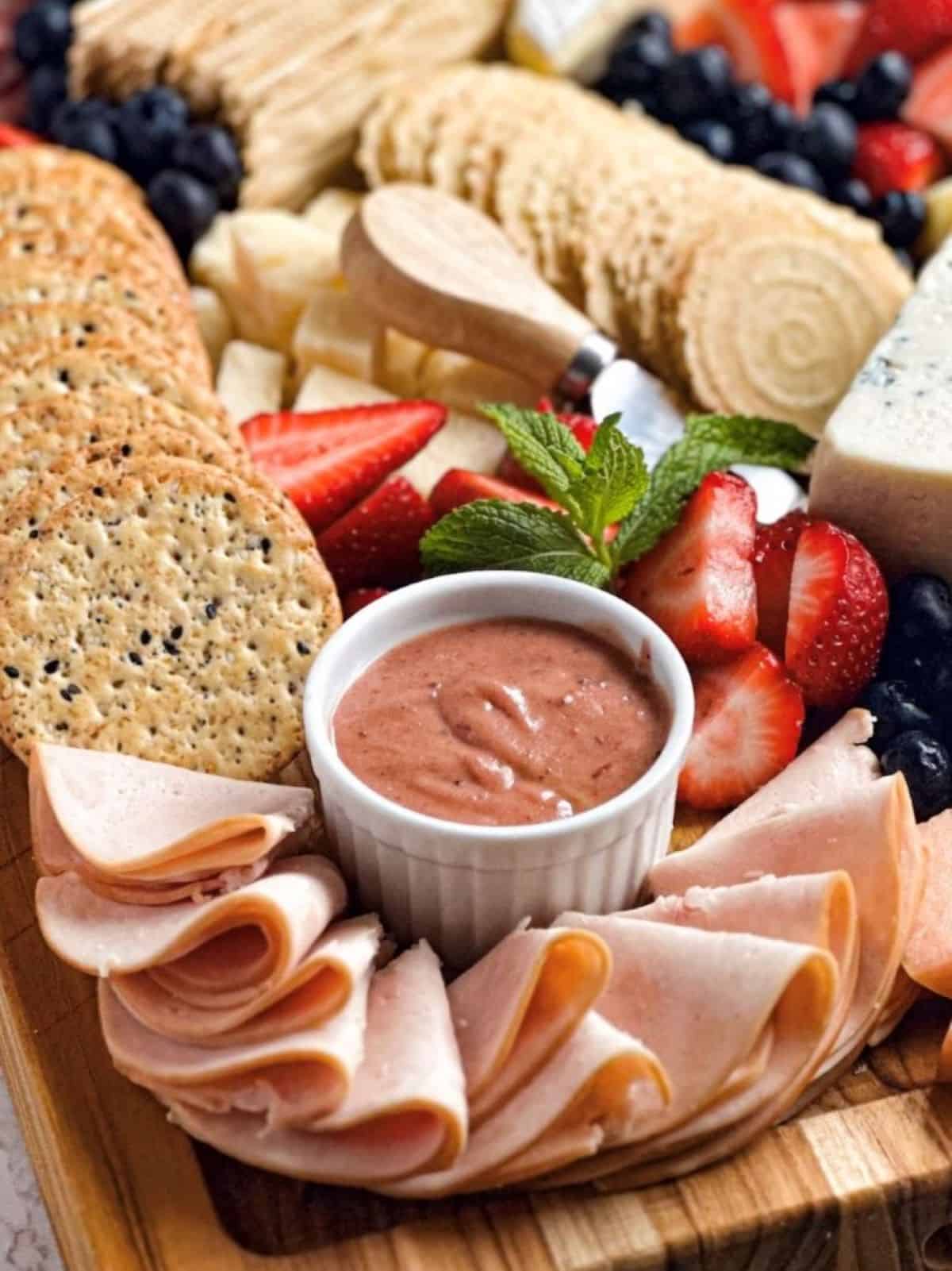 Cranberry mustard in a white ramekin on a charcuterie board with smoked turkey, crackers, cheese, strawberries, and blueberries.