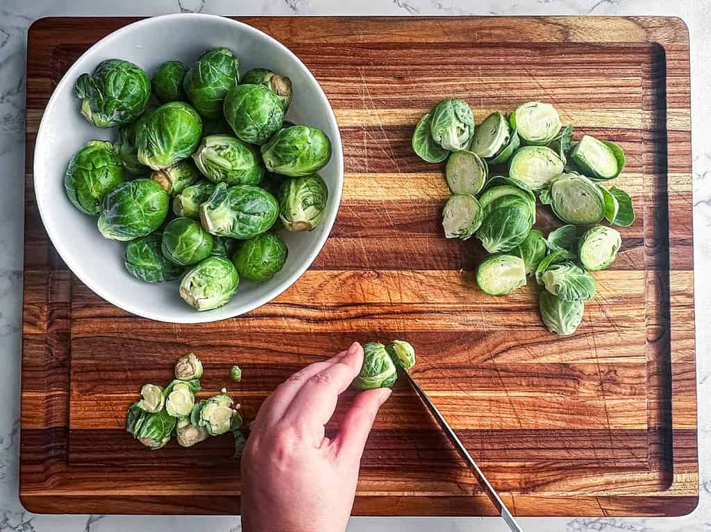 Brussels sprouts being trimmed and halved with a knife on a wooden cutting board.