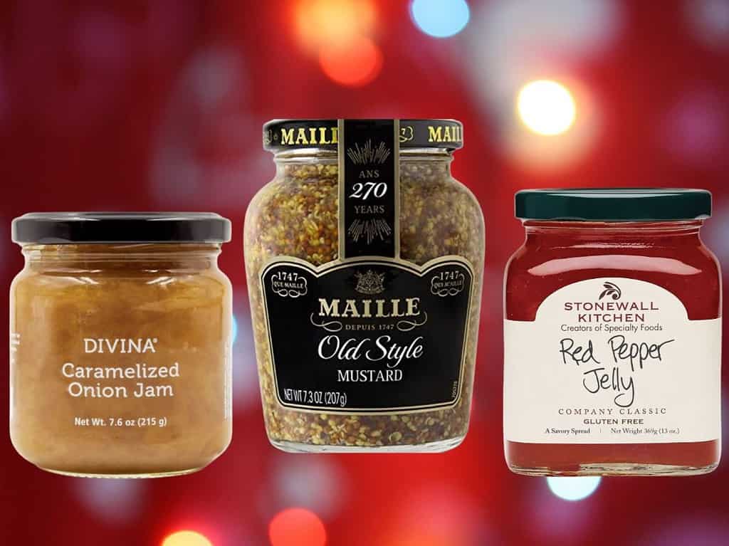 Jars of caramelized onion jam, Maille old style mustard, and red pepper jelly on a red background.
