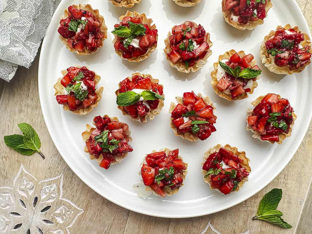 Goat cheese filo cups with balsamic honey strawberries topped with fresh mint on a white plate inside a wooden tray.