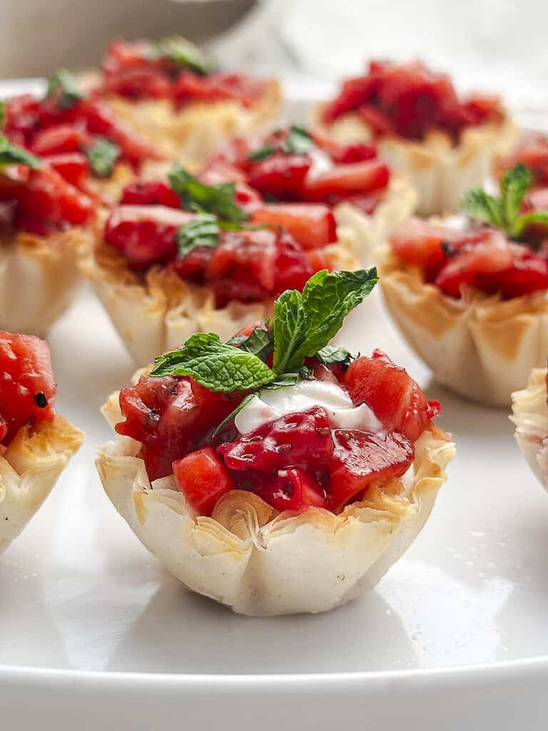 Close up shot of a goat cheese phyllo cup with balsamic strawberries on a white plate with more phyllo cups in the background.