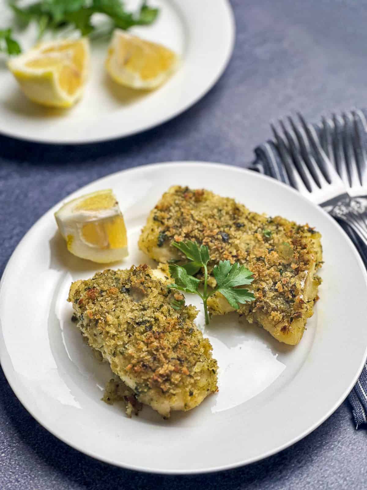 Lemon herb crusted cod on a white plate with slice of lemon and parsley.