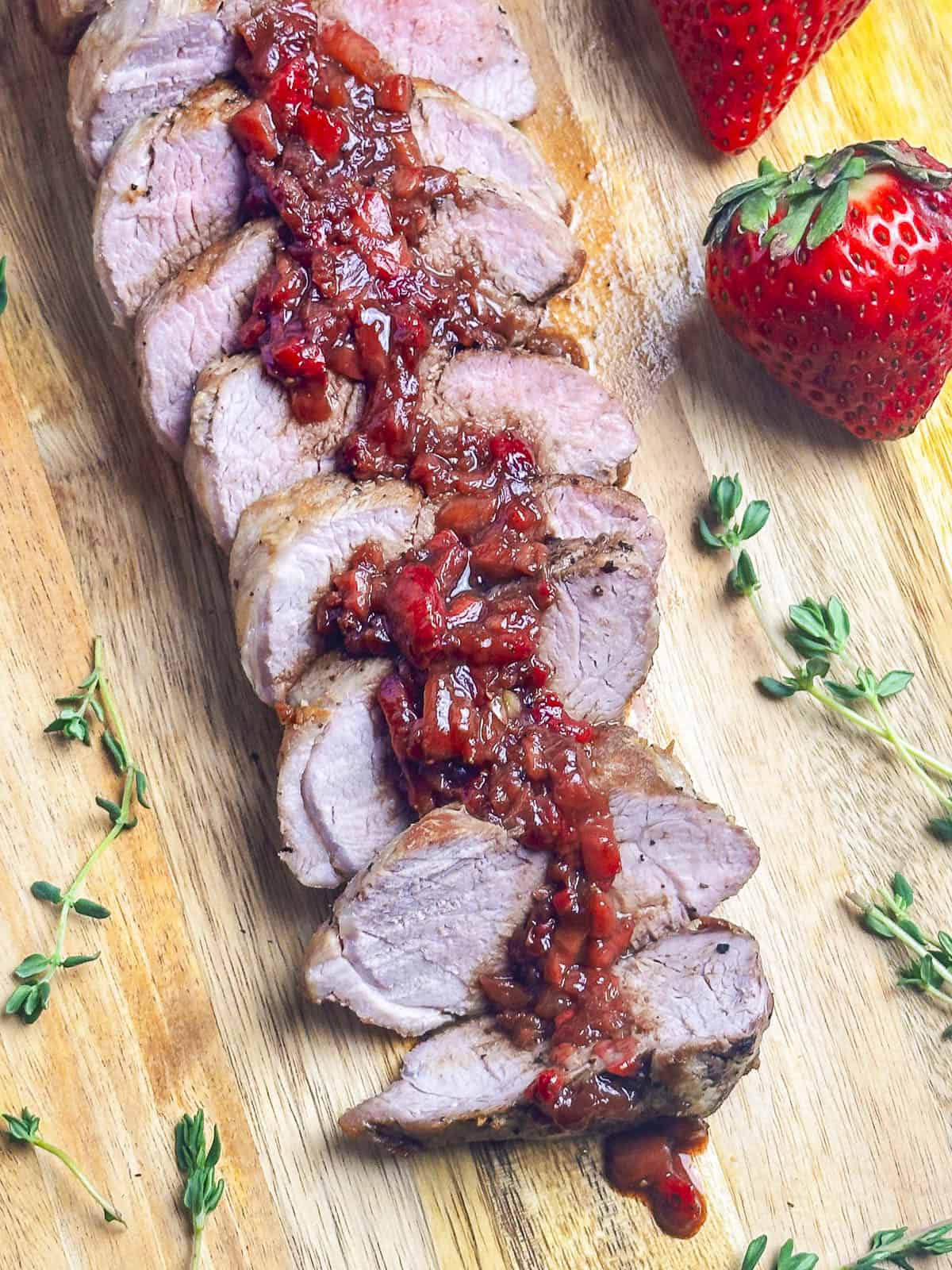 A sliced pork tenderloin with balsamic strawberry sauce on a wooden cutting board with sprigs of thyme surrounding.