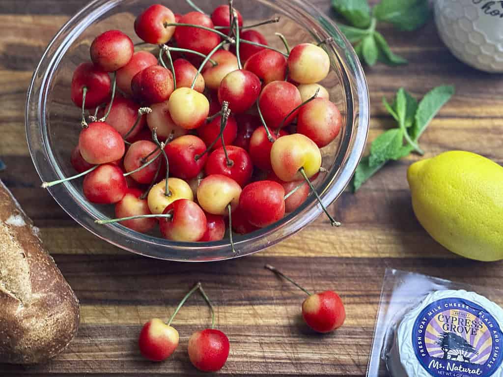 Rainier cherries in a bowl with lemon, goat cheese, bread, and mint on a wooden cutting board.