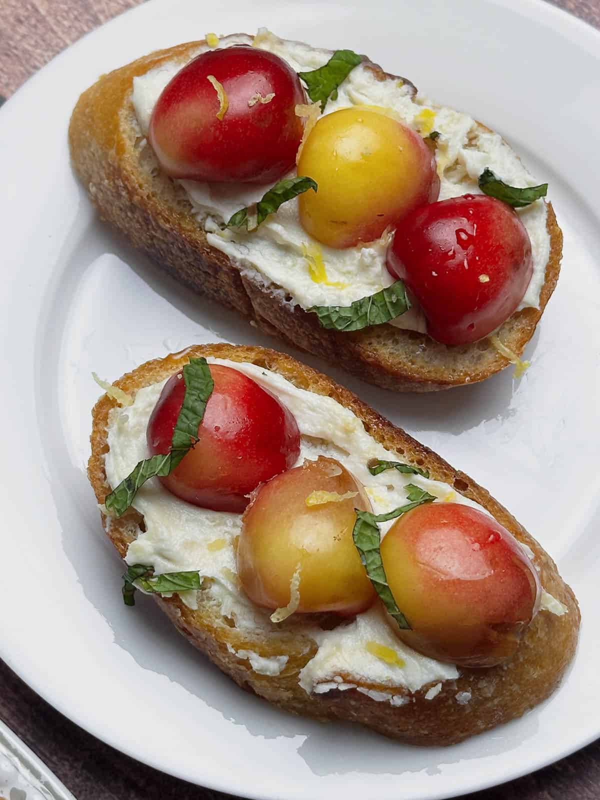 Two goat cheese crostini with Rainier cherries on a white plate.