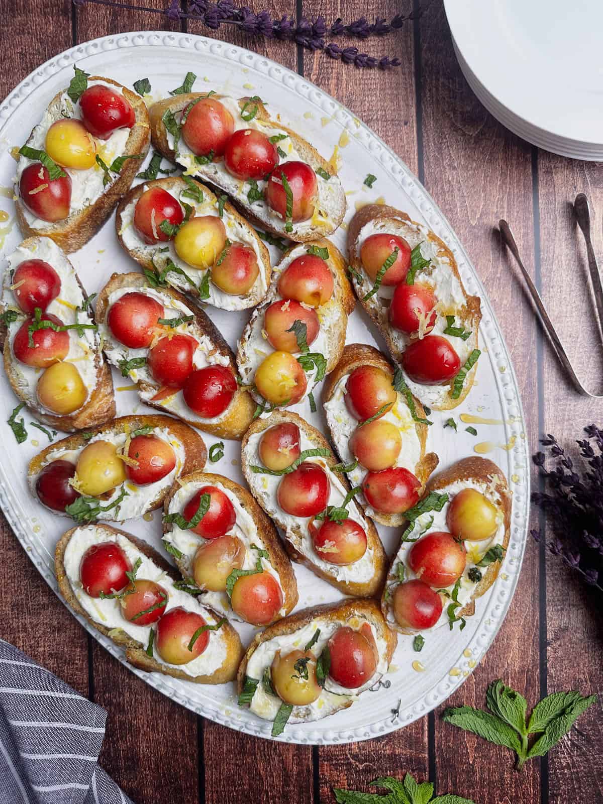 Goat cheese crostini with Rainier cherries on a large white oval serving plate with fresh mint garnish.