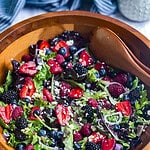 Berries, shallots, goat cheese crumbles, and mixed greens in a wooden salad bowl with a pair of serving spoons.