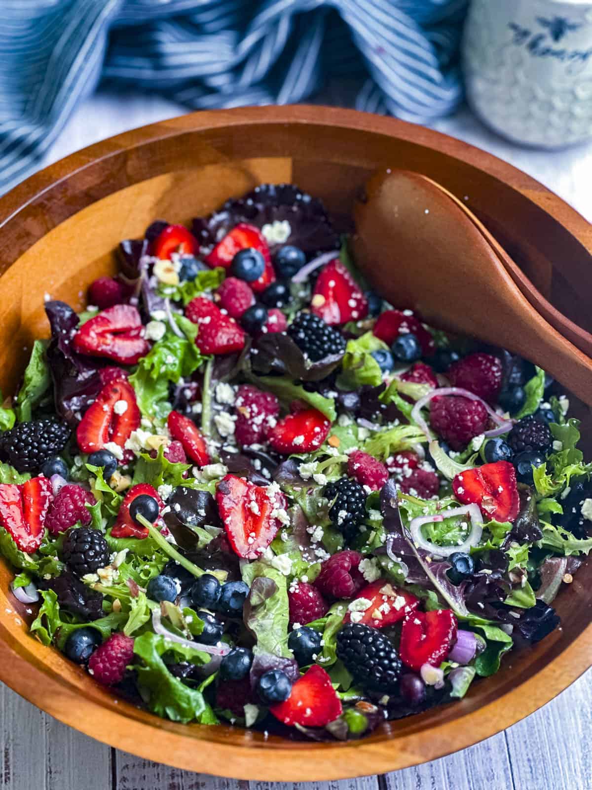 Berries, shallots, goat cheese crumbles, and mixed greens in a wooden salad bowl with a pair of serving spoons.