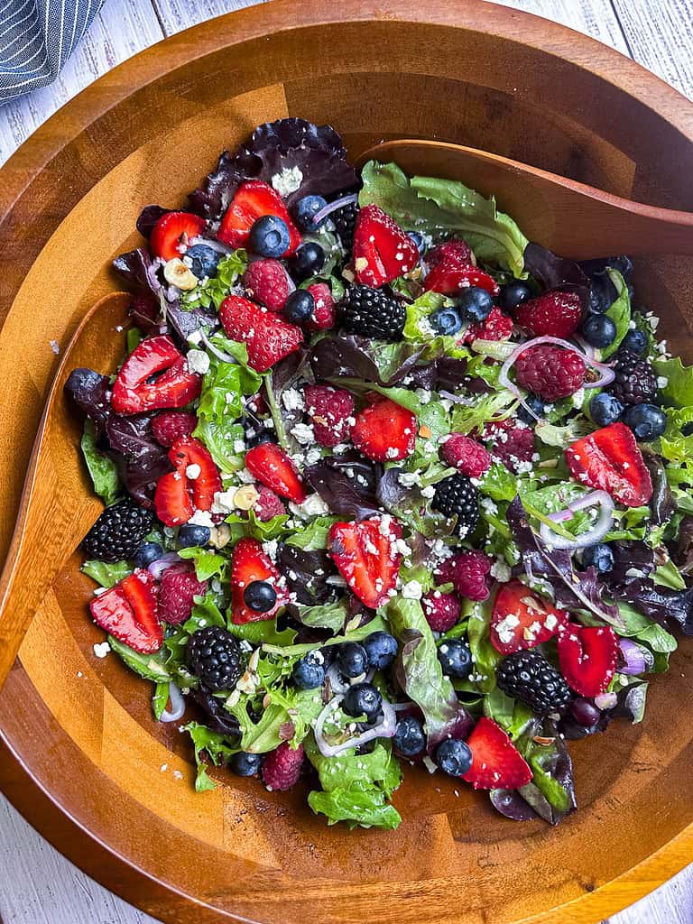 Berries, shallots, goat cheese crumbles, and mixed greens in a wooden salad bowl with a pair of serving spoons.