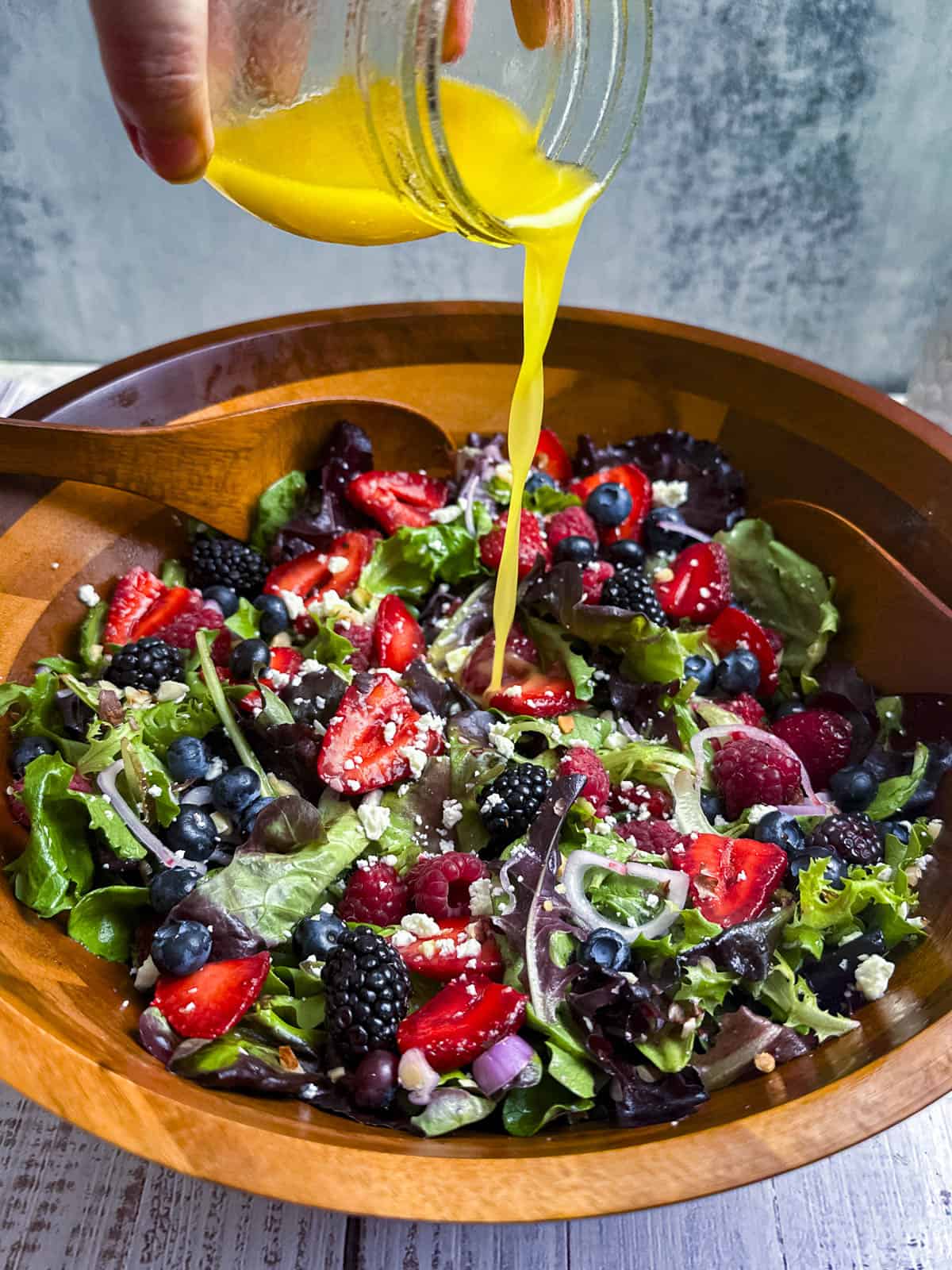 Berries, shallots, goat cheese crumbles, and mixed greens in a wooden salad bowl with a hand pouring dressing over the top.