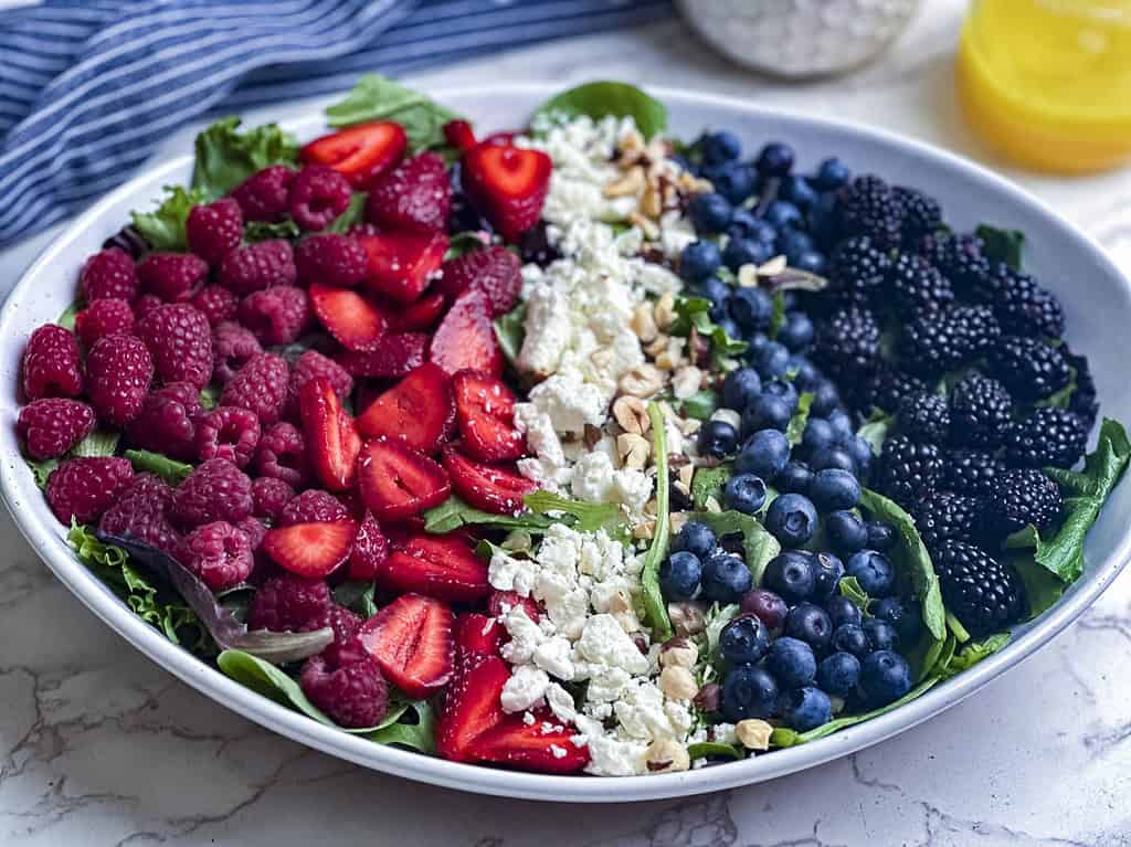 Summer berries, goat cheese, hazelnuts on a bed of mixed greens in a white oval bowl.
