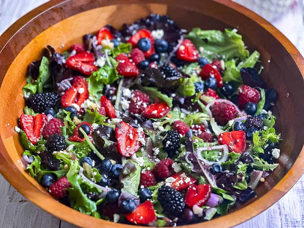 Berries, shallots, goat cheese crumbles, and mixed greens in a wooden salad bowl.