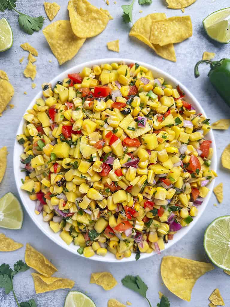 Mango corn salsa in a bowl surrounded by tortilla chips and lime wedges.