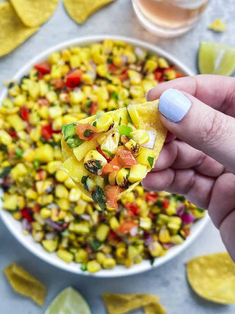Mango corn salsa in a bowl with a hand holding a chip filled with salsa.