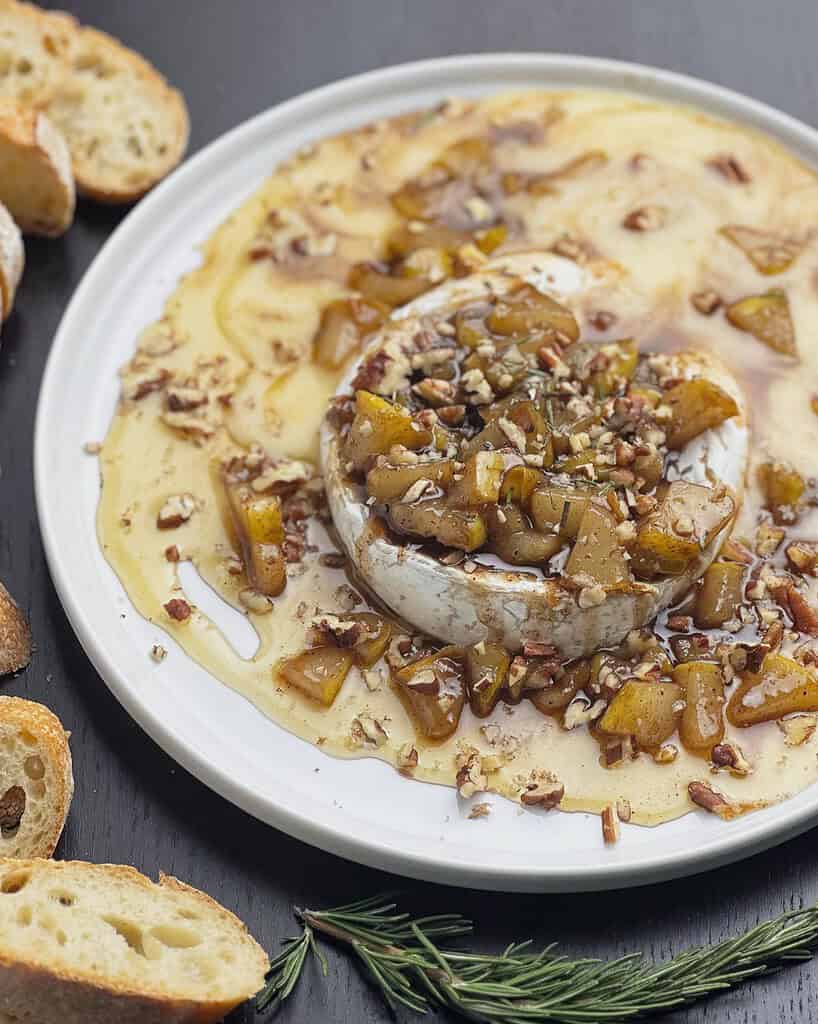Brie with pears, pecans, and honey on a round white plate with sliced baguette on the side.