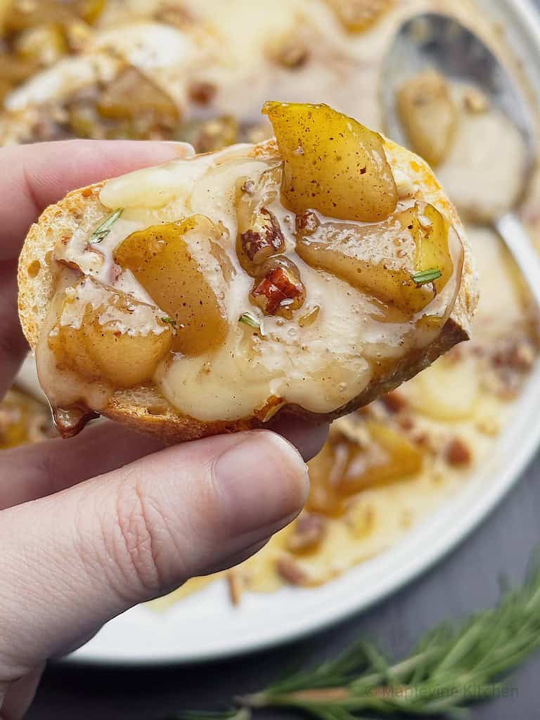 A hand holding a slice of baguette with melted brie cheese and spiced pears.