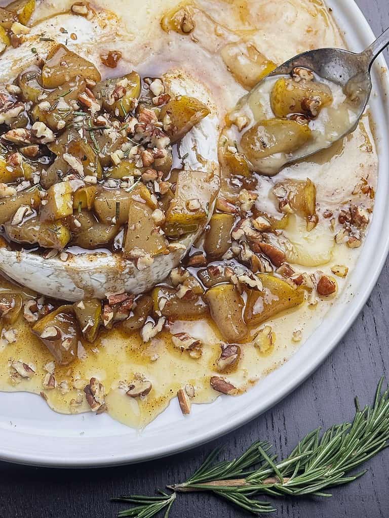 Brie with pears, pecans, and honey on a round white plate with a spoon.