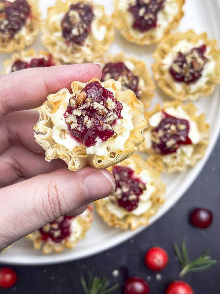 Fingers holding a cranberry tartlet over cranberry tartlets on a round white plate.