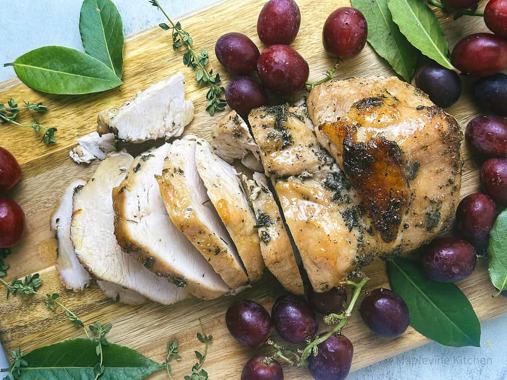 Maple bourbon glazed turkey breast on a wooden serving board with red grapes and fresh green herbs.