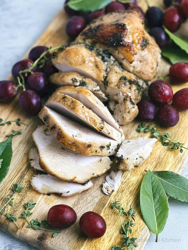 Maple bourbon glazed turkey breast on a wooden serving board with red grapes and fresh green herbs.
