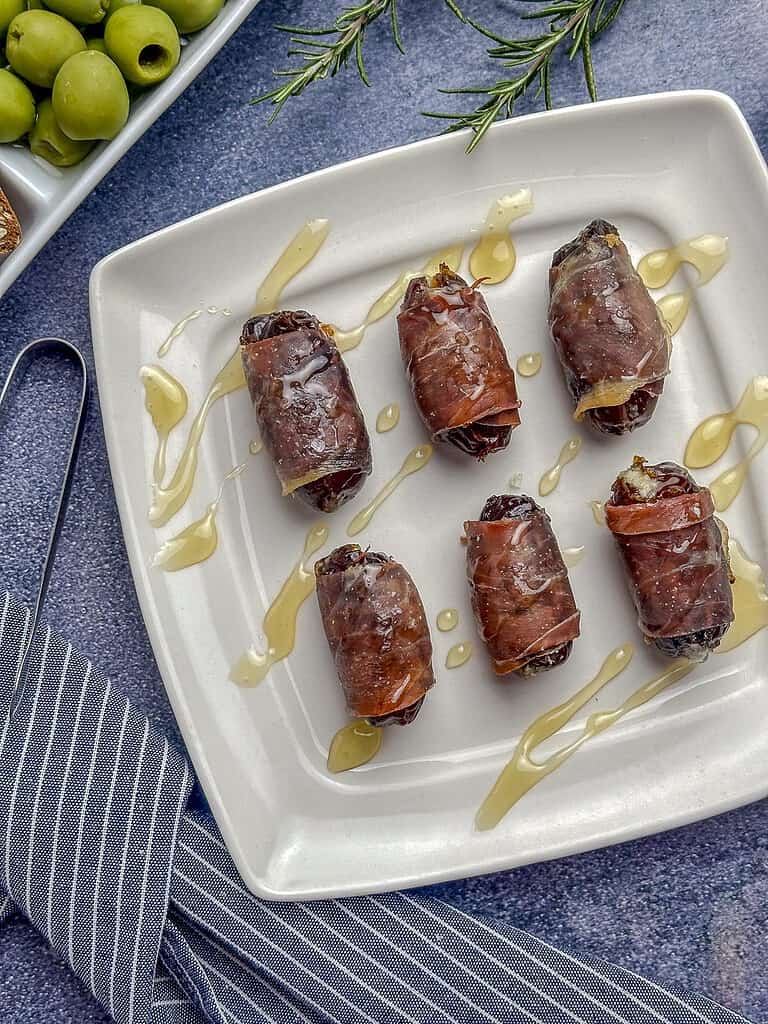 Prosciutto wrapped dates on a plate with honey and a striped napkin on the side.