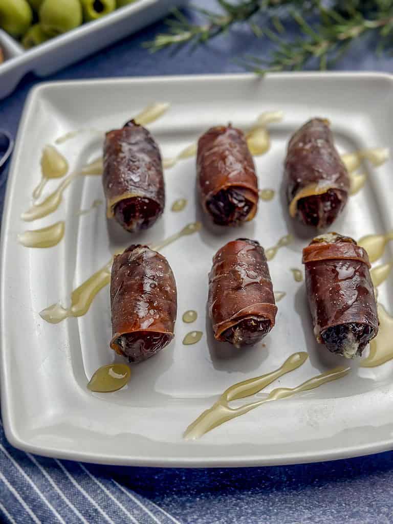 Prosciutto wrapped dates on a white plate with a honey drizzle.
