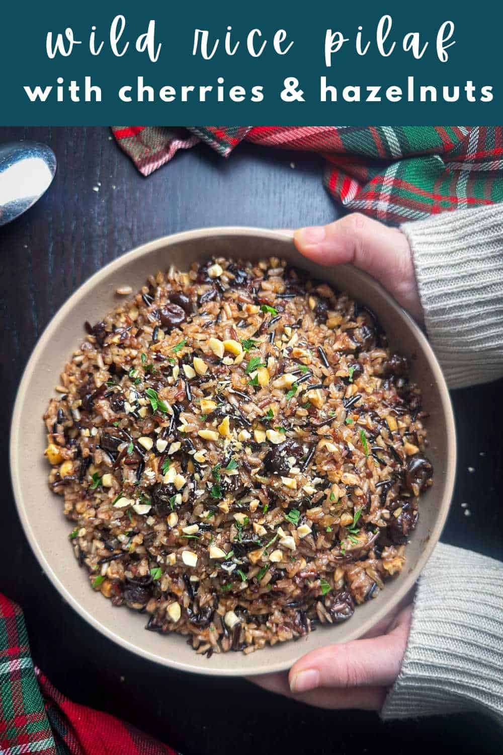 Hands holding a bowl of wild rice pilaf with cherries and hazelnuts.