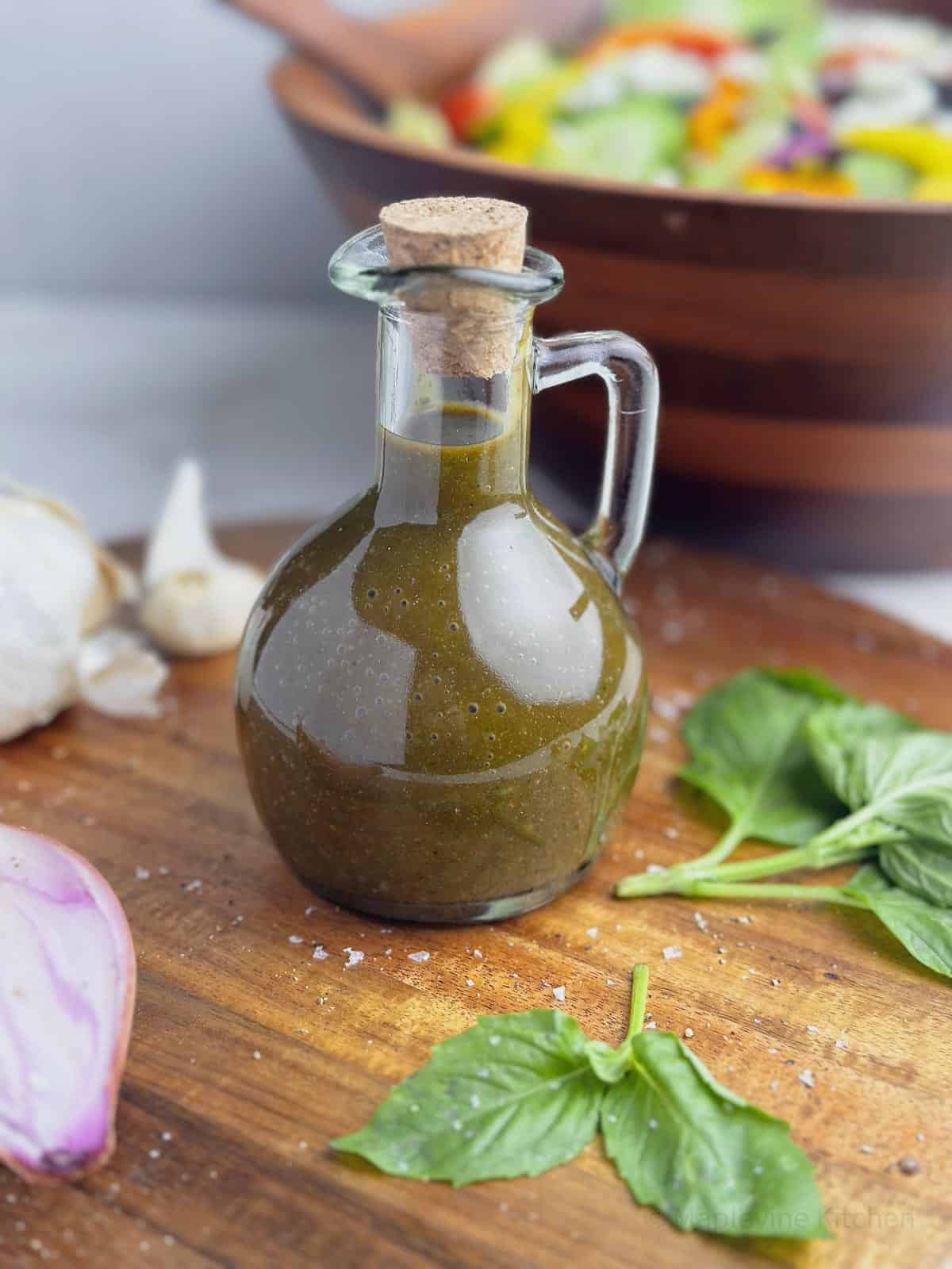 Basil balsamic vinaigrette in a glass carafe with a cork lid surrounded by fresh basil, shallot, and garlic cloves.
